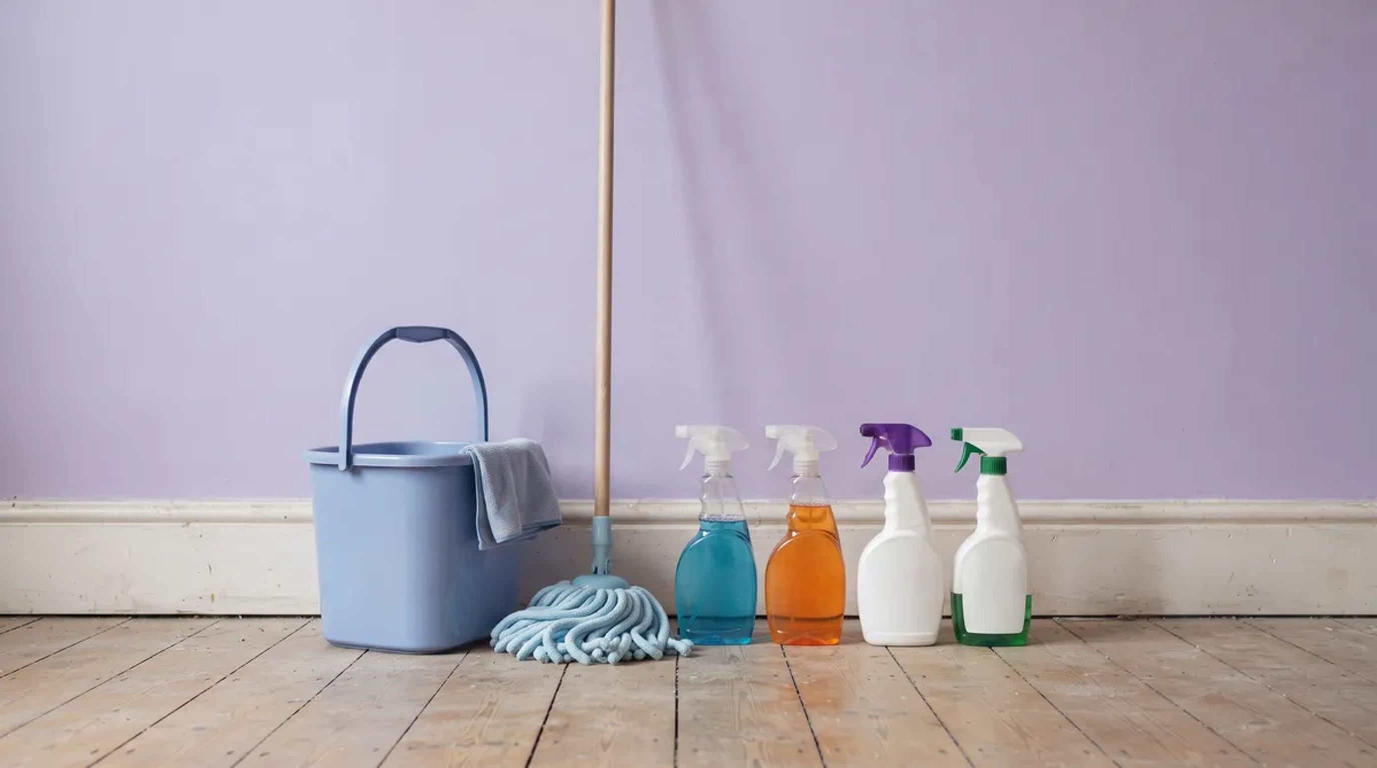 Seau bleu, balai à franges et quatre flacons de produits ménagers posés sur un parquet devant un mur lilas, matériel d'un ménage de location courte durée